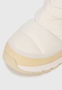 Közelkép egy krém- és bézs színű párnázott sneaker orráról, texturált talppal és varrott szövet részletekkel.