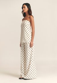 Schulterloser Jumpsuit mit schwarzen Punkten auf cremefarbenem Hintergrund; locker sitzendes, weit geschnittenes Design; leichter Stoff; glatte Textur; minimalistischer Stil.