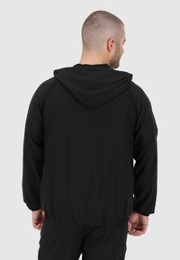 Veste à capuche noire avec une coupe décontractée, taille élastique et panneaux latéraux noirs contrastants. Tissu lisse avec une finition mate.