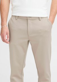 Pantalones beige con una textura suave, corte ajustado y bolsillos laterales discretos. Presentan un cierre de botón y detalles de costura sutiles.