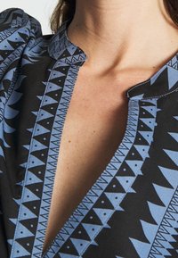 Tissu noir avec un motif géométrique bleu ; présente un col en V et un col texturé. Couture visible le long des bords pour le détail.