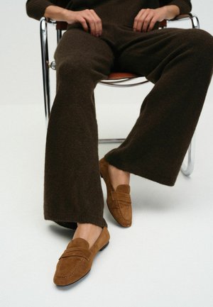 Person sits on chair wearing dark brown knit pants and light brown suede loafers on white floor background.