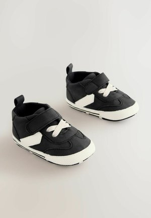 Des chaussures en toile noires avec une semelle blanche, dotées d'une sangle Velcro et de lacets blancs. Le design comprend des coutures discrètes et un accent sur le côté.