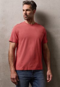 Homme portant un t-shirt rouge uni et un jean bleu, debout contre un mur en béton gris texturé, regardant sur le côté.