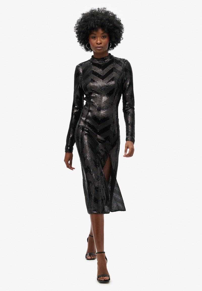 Vestido negro con lentejuelas, de mangas largas, cuello alto, patrón de chevron y abertura alta hasta el muslo. Su silueta ajustada realza la forma del cuerpo.
