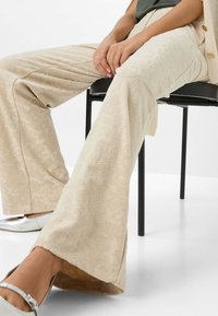 Pantaloni svasati color crema con un motivo floreale testurato, realizzati in tessuto morbido. Abbinati a scarpe silver poggiate su una sedia nera.