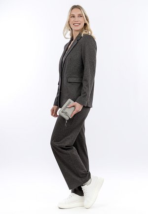 Blondýnka v tmavě šedém obleku a bílých teniskách drží šedou peněženku na zip, usmívá se a dívá se stranou na bílém pozadí.