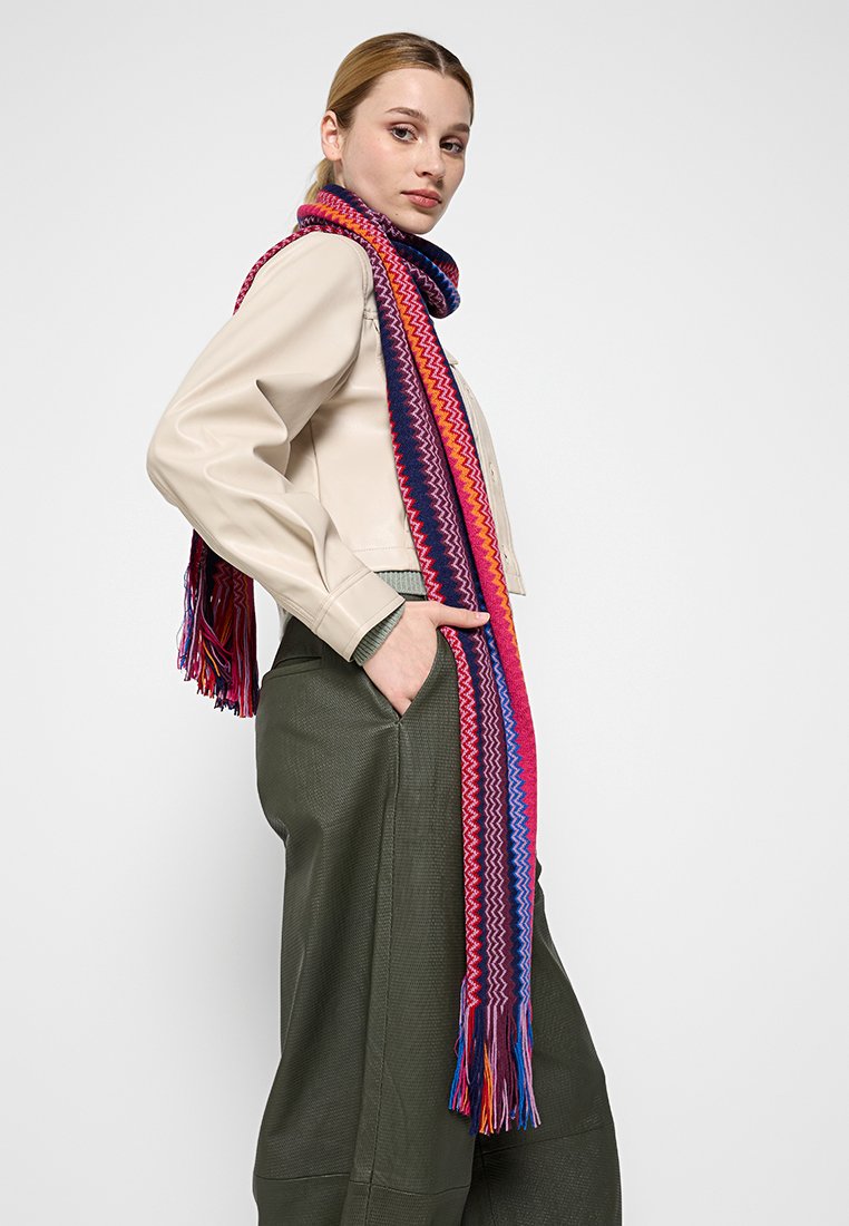 Gewebter Schal in lebhaftem Pink, Lila und Blau mit Zickzackmustern und Fransenkanten, um den Hals eines Modells drapiert.