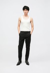 White tank top, black trousers, and black boots. Model stands with hands in pockets, hair is curly and short. Neutral background.