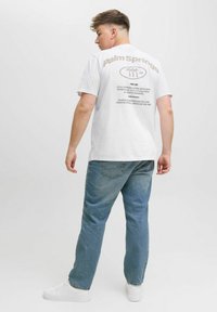 Hvid t-shirt med "Palm Springs" trykt i brunt på ryggen, parret med lyseblå jeans og hvide sneakers. Simpelt, afslappet design.
