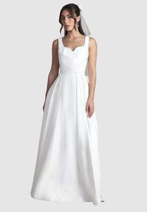 Donna in un abito da sposa bianco senza maniche con corpetto aderente, gonna fluente e velo legato con un fiocco nei suoi capelli scuri.