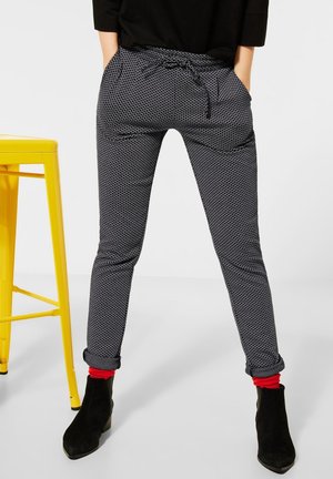 Persoon draagt zwart-witte broek met omgeslagen boorden, rode sokken, zwarte enkellaarsjes en een zwart topje, staand naast een gele kruk.