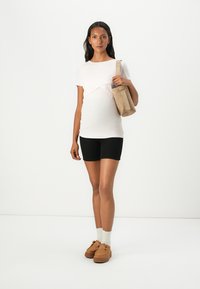Tricou de maternitate de culoare roz deschis, cu un design ajustat și un overlay curbat; asortat cu pantaloni scurți negri și pantofi slip-on maro, ținând o geantă bej.