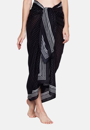 Person trägt einen schwarzen Sarong mit weißen Punkten und gestreiftem Rand, um die Taille gebunden, steht barfuß vor einem einfarbigen Hintergrund.