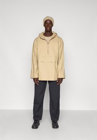 Beiger Pullover mit Halbzipp, großer Kapuze und vorderer Tasche, kombiniert mit dunkelgrauen Cargo-Hosen und schwarzen Schuhen. Einheitliche Farben, keine Muster.