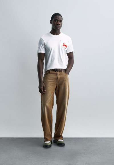 Άνδρας που στέκεται σε λευκό τοίχο φορώντας λευκό μπλουζάκι με κόκκινο άλογο στην τσέπη, καφέ παντελόνι με φαρδιά πόδια, σκούρα ζώνη και δίχρωμα παπούτσια.