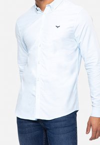 Camisa de botón de color azul claro, hecha de algodón, con mangas largas, que presenta un collar clásico y un pequeño logotipo bordado en el pecho.