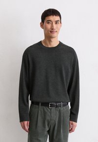 Dunkelgrüner Langarmpullover aus weichem, strukturiertem Material, kombiniert mit hellgrünen Cordhosen und einem schwarzen Gurt.