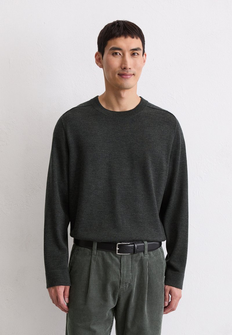 Un maglione lungo verde scuro a maniche lunghe realizzato in un materiale morbido e testurizzato, abbinato a pantaloni in corduroy verde chiaro e una cintura nera.