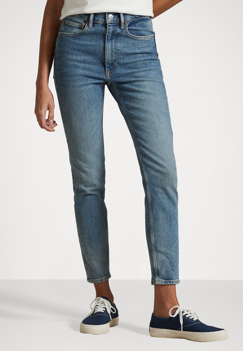 Pessoa vestindo jeans azuis slim de cintura média e ténis de lona azul marinho com atacadores brancos, em pé sobre um chão branco contra um fundo branco.