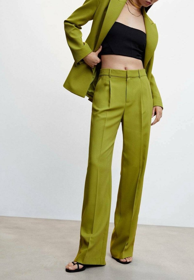Vrouw draagt olijfgroene op maat gemaakte broek en bijpassende blazer, zwart strapless crop top, zwarte sandalen en gouden kettingen.