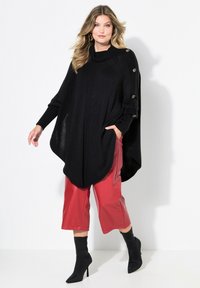 Poncho en laine noir avec col montant, featuring des accents côtelés et des boutons dorés aux manches. Associé à un pantalon en cuir rouge et des bottes noires.
