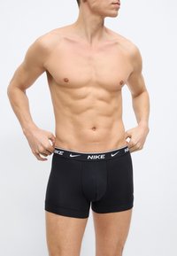 Άνδρας χωρίς πουκάμισο που ρυθμίζει μαύρα μποξέρ Nike με λανσαρισμένη ζώνη μέσης, ορατός καλογυμνασμένος κορμός και μυώδη χέρια σε απλό φόντο.