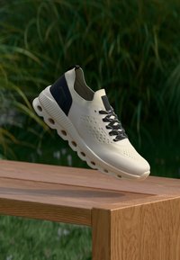 Chaussure de course légère coloris crème avec une tige texturée, un accent talon bleu foncé et des lacets noirs. Dispose d'une semelle coussinée avec des découpes circulaires.