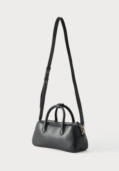 Μαύρη δερμάτινη ορθογώνια τσάντα με δύο κοντές λαβές και έναν μακρύ ρυθμιζόμενο ιμάντα ώμου, με φερμουάρ, που στέκεται πάνω σε λευκή επιφάνεια.