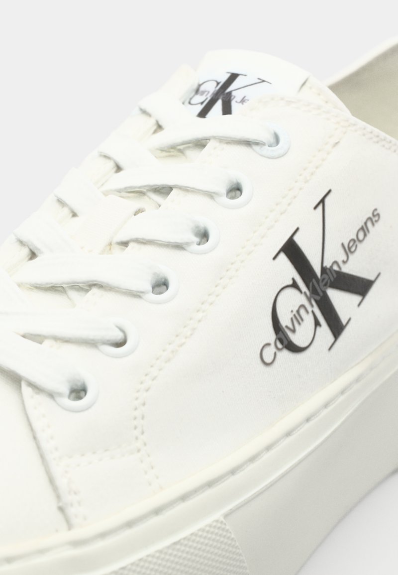 Baskets en toile blanches avec un bout arrondi, un système de laçage et une semelle épaisse. Présente un logo "CK" noir et le texte "Calvin Klein Jeans" imprimé.