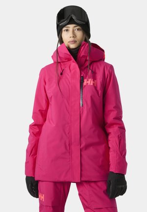Joven mujer con chaqueta y pantalones de esquí de color rosa brillante, guantes negros, gorro negro y gafas de esquí en la cabeza, mirando hacia adelante.
