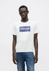 Λευκό βαμβακερό T-shirt με μπλε ορθογώνιο λογότυπο που αναγράφει "JACK&JONES" σε λευκά και κόκκινα γράμματα. Φοριέται με μπλε τζιν παντελόνι.