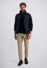 Marineblauwe puffer vest met hoge kraag, ritssluitingen aan de voorkant en een textuur navy trui eronder, gecombineerd met lichtbeige broeken en donkere schoenen.