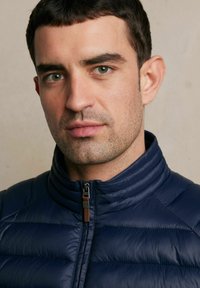 Granatowa kurtka puchowa z wysokim kołnierzem, z zamkiem błyskawicznym oraz skórzanym detalem na uchwycie zamka. Gładka, pikowana tekstura.