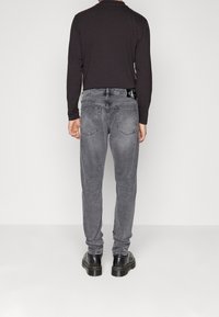 Jean en denim gris à coupe droite, avec poches arrière, détails de couture et un écusson de marque sur la taille. Porté avec des bottes noires.