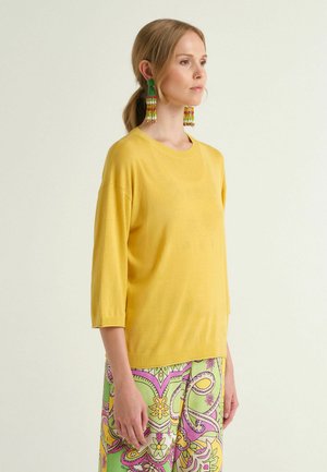 Donna con una maglia gialla a maniche tre quarti, pantaloni a fantasia verdi, rosa e gialli, e lunghe orecchini infilati multicolori, in piedi di profilo.