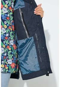 Persona che tiene aperta una giacca blu navy mostrando la tasca interna con cerniera e fodera azzurro chiaro, indossando un cappotto con motivo floreale.