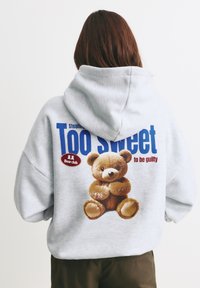 Szary, oversize'owy hoodie z dużą grafiką brązowego misia pluszowego oraz napisem "Zbyt słodki, by być winnym" w niebieskim i czerwonym kolorze. Miękka tekstura materiału.