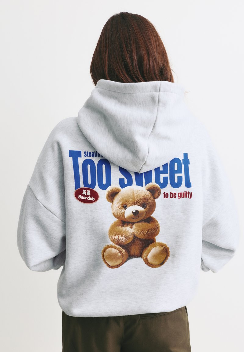 Szary, oversize'owy hoodie z dużą grafiką brązowego misia pluszowego oraz napisem "Zbyt słodki, by być winnym" w niebieskim i czerwonym kolorze. Miękka tekstura materiału.