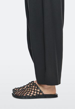 Personne portant un pantalon noir à jambes larges et des sandales tissées noires sans bride, montrant le pied nu à travers le design en filet.