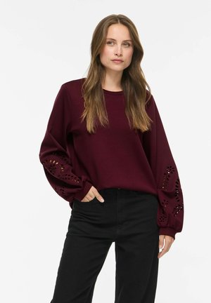 Vrouw met lang bruin haar die een bordeauxrode top met lange mouwen draagt met laser gesneden bloemmotieven op de mouwen en een zwarte broek, staand met één hand in de zak.