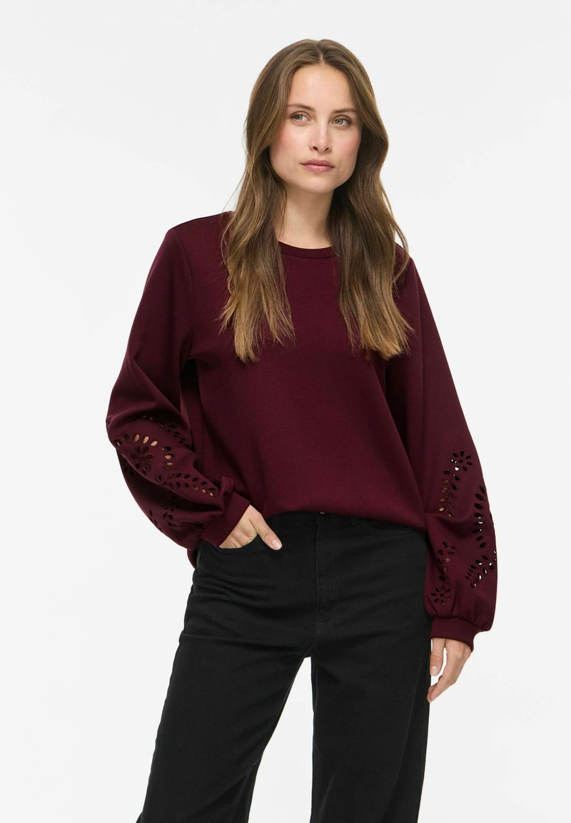 Femme aux longs cheveux bruns portant un haut bordeaux à manches longues avec des motifs floraux découpés au laser sur les manches et un pantalon noir, debout avec une main dans la poche.