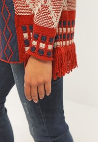 Main reposant sur le côté, portant un jean bleu et un pull en tricot à motifs rouges, blancs et bleus avec des franges au bord de la manche.