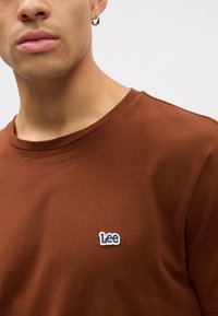 Barna pamut póló kerek nyakkal és rövid ujjakkal, fehér és kék hímzett "Lee" logóval a mellkason.