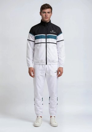 Ung mann står kledd i en hvit treningsdress med svarte og turkise striper og hvite joggesko mot en enkel, lys bakgrunn.