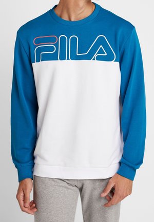 Mand iført blå og hvid FILA sweatshirt med lange ærmer og lysegrå bukser mod en ensfarvet baggrund.