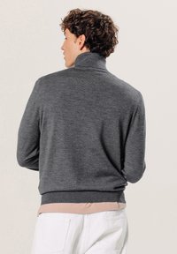 Grijze gebreide trui met hoge kraag en geribde manchetten, gedragen over een lichtroze gelaagd shirt met een gladde structuur.