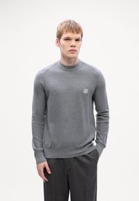 Pulover gri tricotat cu guler rotund, model texturat și o etichetă logo subtilă pe pieptul stâng, combinat cu pantaloni cu model.