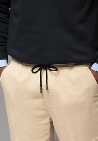 Beige Joggingshosen mit schwarzem Kordelzug, mit einer glatten Textur und elastischem Bund, kombiniert mit einem dunklen Pullover mit hellen Kragen.