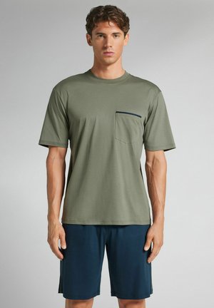 Hombre con una camiseta de manga corta verde oliva con bolsillo en el pecho y pantalones cortos azul marino, de pie contra un fondo gris liso.